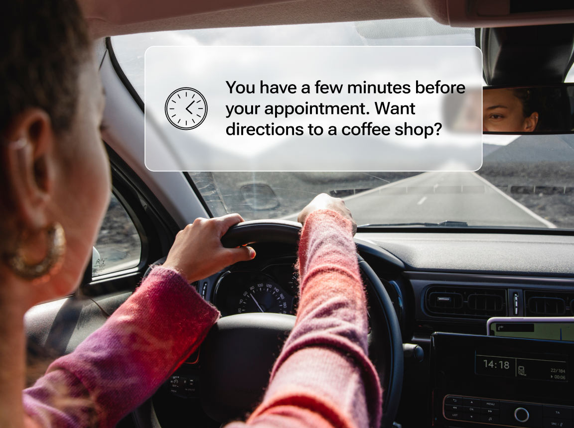 A view from the backseat of a driver with her hands on the wheel, and a notification that says: “You have a few minutes before your appointment. Want directions to a coffee shop?”